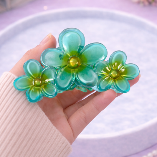 Hand holding a green floral hair clip on a light purple surface with flowers in the background