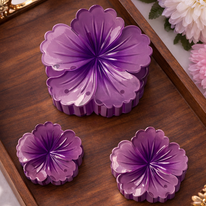 Three purple floral-shaped objects on a wooden tray with gold accents, surrounded by pink and white flowers.