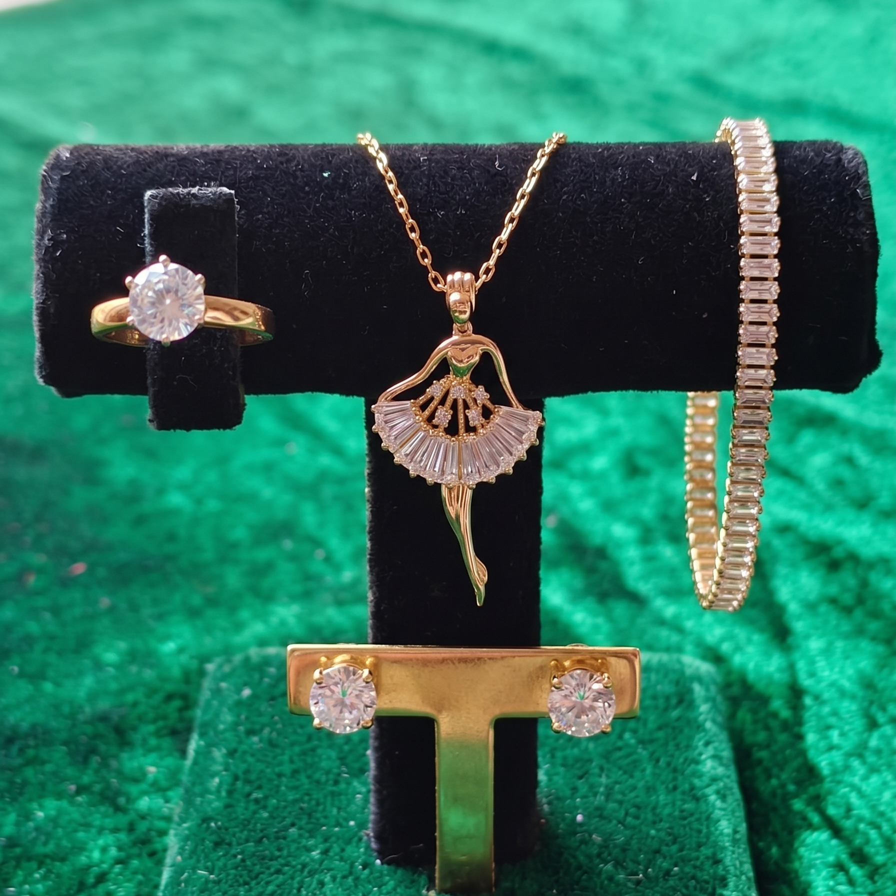 Gold jewelry set including a ring, necklace, and bracelet on a black display stand against a green background.