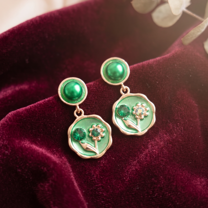 Green floral earrings on a burgundy velvet surface with a blurred natural background