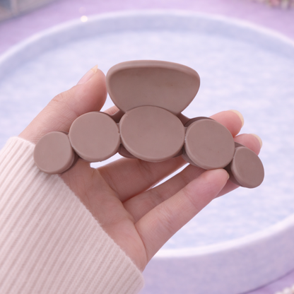 Hand holding a set of beige hair clips on a light purple surface with a book and flowers in the background.