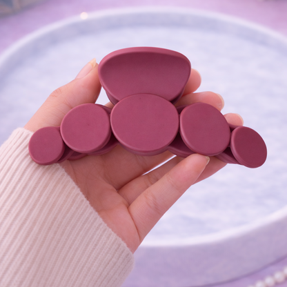 Hand holding a set of pink hair clips on a light purple surface with a book and flowers in the background.
