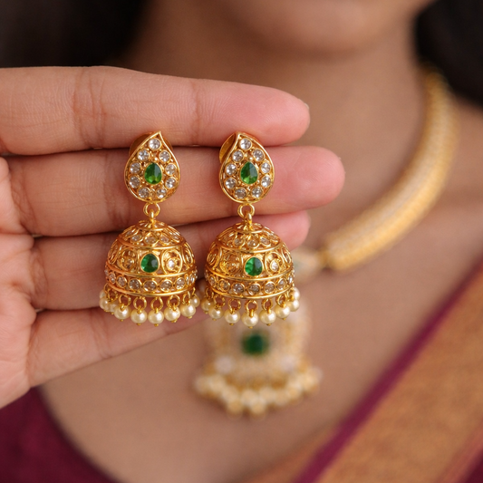 Gold earrings with green gemstones held by a person wearing a maroon saree.