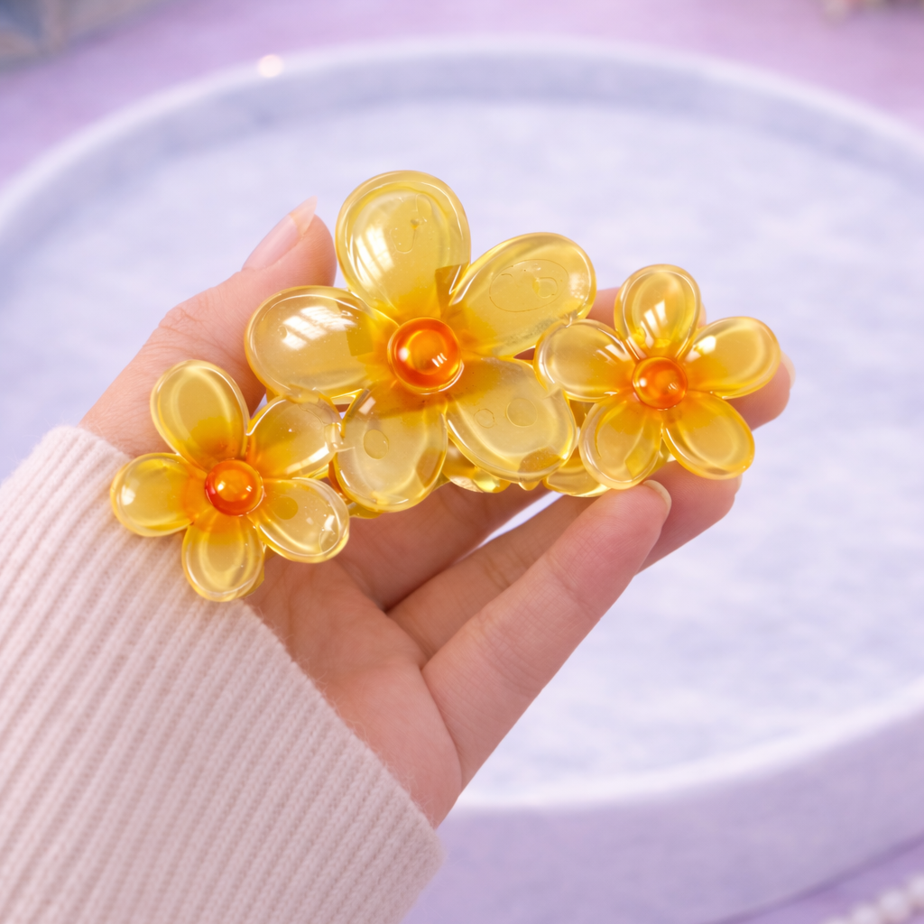 Hand holding a yellow flower-shaped hair clip on a light purple surface with a book and flowers in the background.