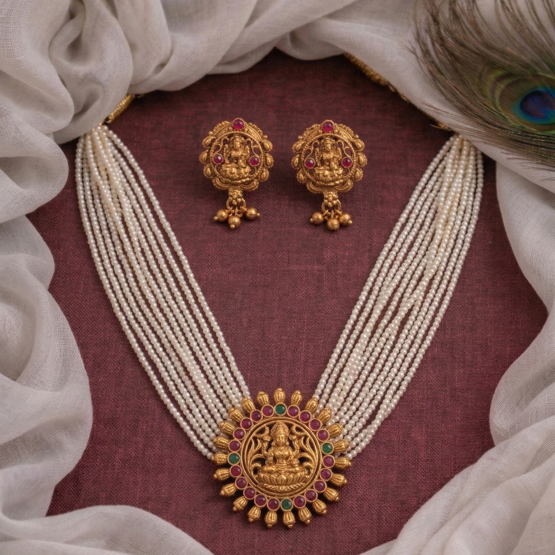 Gold necklace and earrings with red and green stones on a maroon fabric background