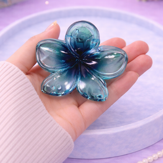 Hand holding a blue flower-shaped object on a purple surface with a pearl necklace.