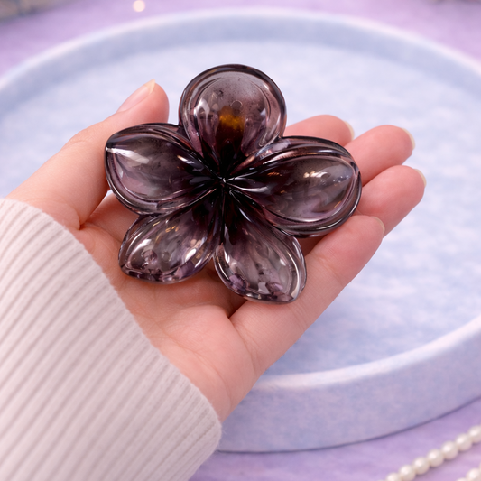 Hand holding a purple flower-shaped object on a light purple surface with a book and flowers in the background.